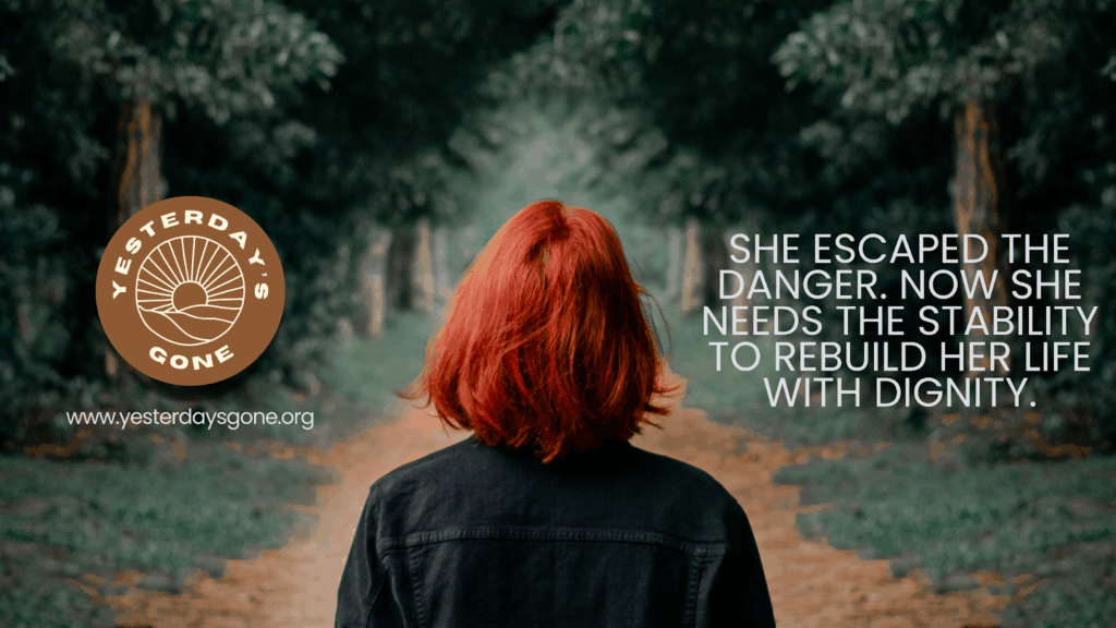 A woman with red hair stands with her back to the camera on a wooded path, symbolizing a journey toward safety and stability. The Yesterday’s Gone logo appears on the left, along with the website. On the right, the text reads: ‘She escaped the danger. Now she needs the stability to rebuild her life with dignity.